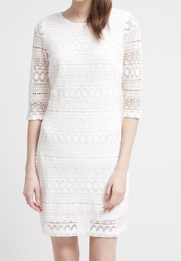 Femme portant une robe en crochet blanche, longueur genou, à manches trois-quarts, sur un fond uni.