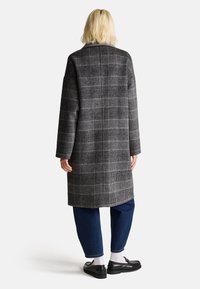 Cappotto lungo a quadri grigio con accenti neri e beige, realizzato in tessuto testurizzato. Presenta una linea dritta, maniche lunghe e tasche laterali.