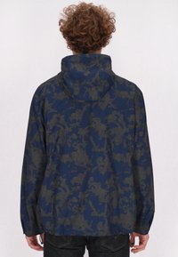 Jachetă cu glugă de culoare bleumarin, cu un model geometric gri, având manșete elastice și un tiv cu șnur. Material neted, ușor.