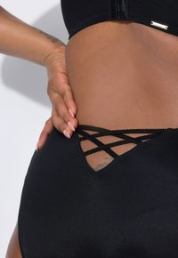 Zwarte zwemkleding met een gestructureerde afwerking, met een V-vormige uitsnijding en kruislings gebonden banden, en een subtiele logo-accent. Zachte stof en delicate details.