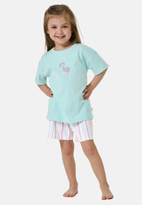 Camiseta de algodón azul claro con un gráfico de flamenco rosa, combinada con pantalones cortos a rayas en tonos pastel. El modelo está de pie con las manos en las caderas, descalzo.