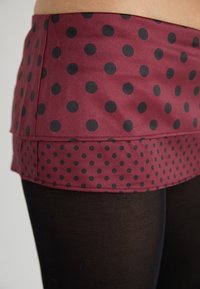 Tissu bordeaux avec des pois noirs, comprenant deux couches : un haut uni et un ourlet volanté à pois. Associé à des leggings noirs.