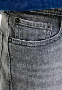 Gros plan d'une petite poche avant sur un jean en denim gris avec des rivets noirs, partiellement recouverte d'un tissu bleu en haut.