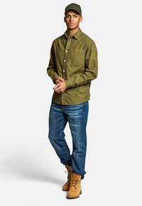Camicia a maniche lunghe verde oliva, jeans in denim azzurro chiaro, stivali da lavoro beige e berretto verde con logo. La camicia ha una tasca sul petto e polsini con bottone.