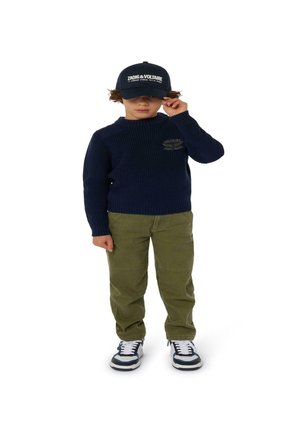 Marine ribbed trui, olijfgroene broek, zwarte pet met witte letters, en witte sneakers met marineblauwe accenten op een kind dat tegen een witte achtergrond staat.