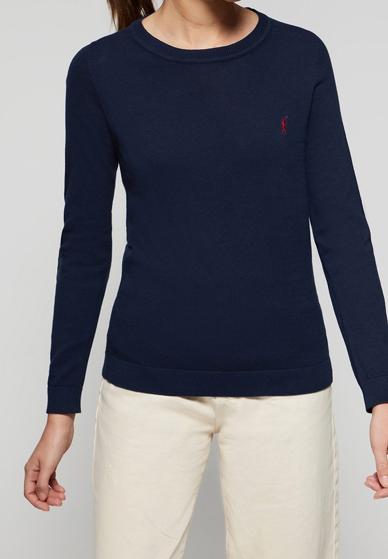 Femme portant un pull ajusté à manches longues bleu marine avec un logo rouge sur la poitrine et un pantalon beige clair, sur un fond gris uni.