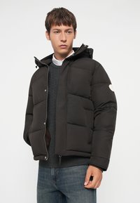 Sort puffer jakke med hætte, med quiltede sektioner, lynlås lukning og et logo patch på venstre ærme. Sammen med en mørk sweater.