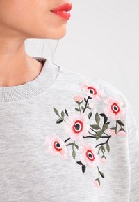 Sweatshirt grigio con ricami floreali dettagliati in rosa e rosso, con foglie verdi, attorno al collo e alla zona superiore delle spalle.