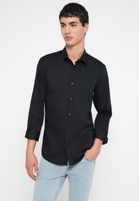 Fekete hosszú ujjú ing sima anyagból, klasszikus gallérral és gombos elülső résszel, felhajtott mandzsettákkal és testhez simuló szabással.