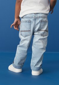 Jean en denim bleu clair avec une coupe décontractée, ourlets retroussés et deux poches arrière. Associé à des baskets blanches sur un fond bleu.