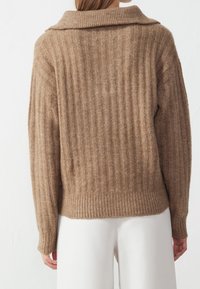 Femme portant un pull en laine marron côtelé avec un large col, vue de dos, associée à un pantalon blanc.