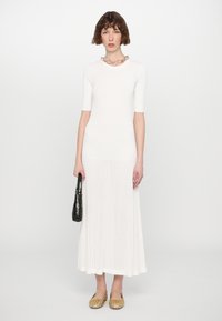 Robe en tricot côtelé blanche avec des manches courtes, une longue jupe et un collier en chaîne. La mannequin tient un sac à main noir et porte des ballerines dorées tressées.