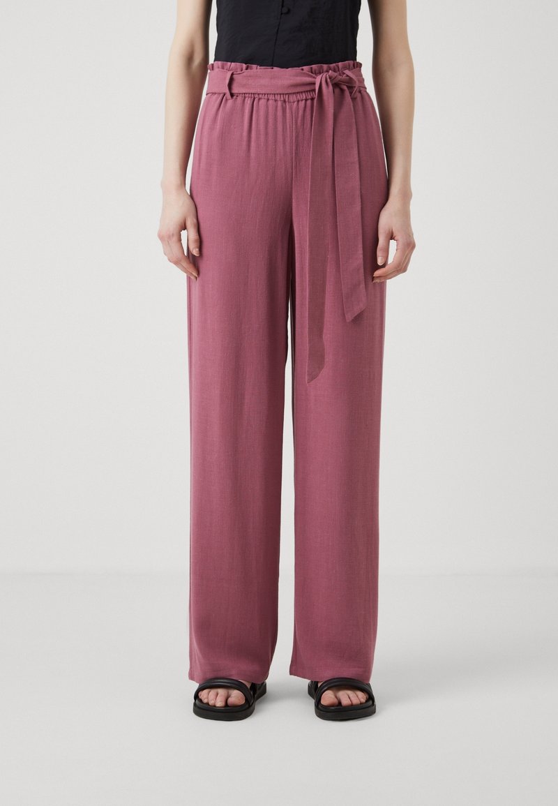 Femme portant un pantalon rose ample taille haute avec ceinture nouée, un haut noir sans manches et des sandales noires à bout ouvert sur un fond uni.