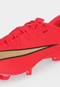 Φωτεινό κόκκινο ποδοσφαιρικό παπούτσι Nike με χρυσό λογότυπο Swoosh, κορδόνια και καλουπωμένα τακούνια για παιχνίδι σε σκληρό έδαφος.