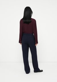 Woman with straight black hair wearing a burgundy long-sleeve shirt tucked into navy tailored pants and black shoes, standing facing a white wall.