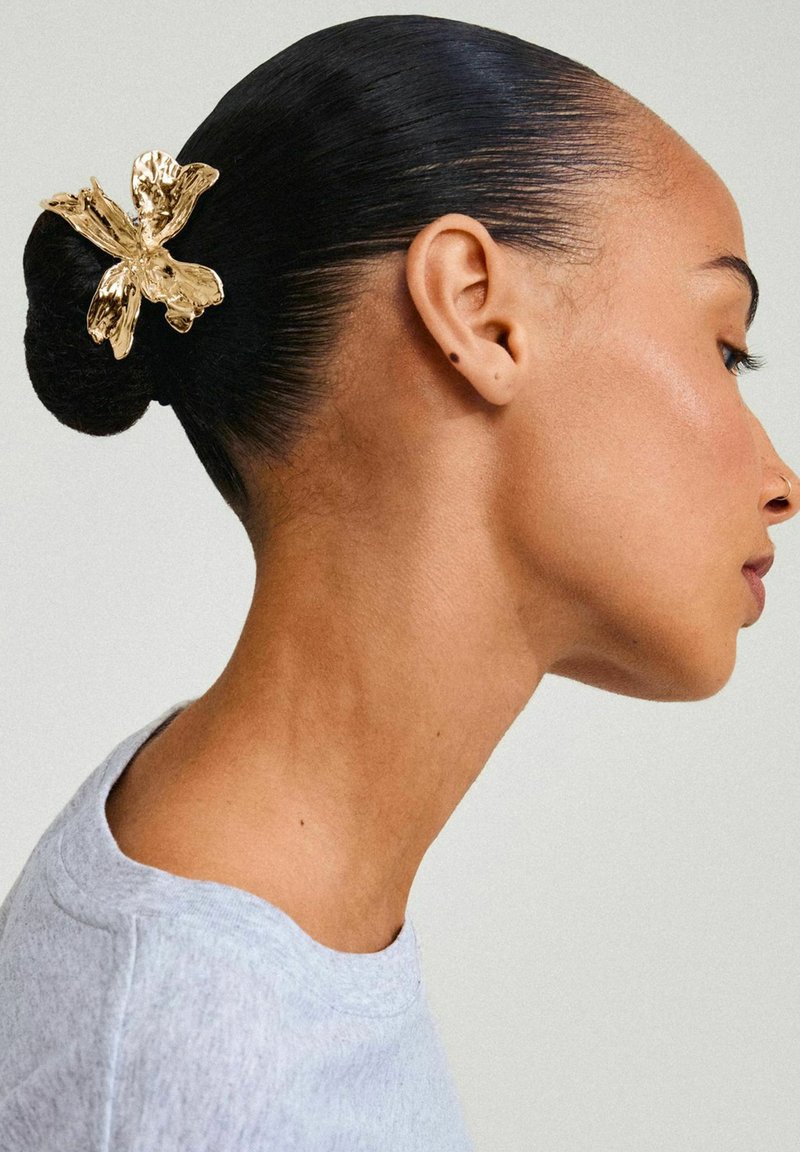 Goldene Blumenhaarspange, die in einem eleganten schwarzen Dutt platziert ist; das Material hat eine strukturierte, metallische Oberfläche. Graues Rundhalstopp ist teilweise sichtbar.