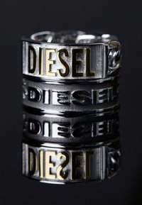 Anillo de metal plateado y negro con la palabra "DIESEL" en letras doradas. El anillo tiene una superficie texturizada y un acabado brillante.