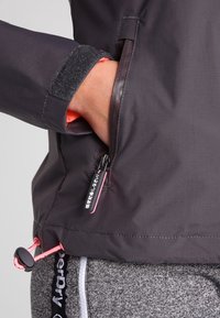 Ruka zasunutá do kapsy na zip na tmavě šedé bundě s růžovými akcenty a nastavitelným šňůrovým lemem přes šedé texturované kalhoty.