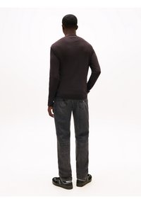 Man standing with back to camera wearing dark long-sleeve shirt, faded black jeans, and black sneakers on white background.