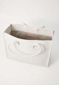 Sac fourre-tout en cuir blanc avec des poignées incurvées, surface texturée, logo en relief, et une doublure intérieure beige avec une petite étiquette.