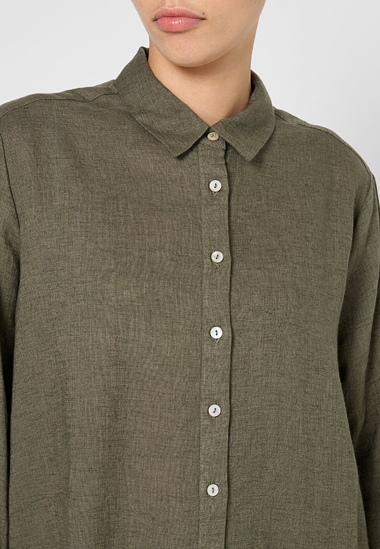 Chemise olive, à boutons, fabriquée dans un tissu texturé. Dotée d'un col et de six boutons de couleur claire sur le devant. Manches longues.