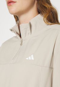 Femme portant un haut beige à fermeture éclair quart de zip avec un col haut et un petit logo blanc triangulaire sur la poitrine, lèvres et partie des cheveux visibles.