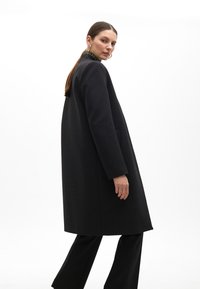 Black wool coat with a straight cut, side pockets, and a high collar, worn over black trousers. Smooth texture, minimalistic design.