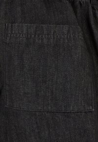 Tessuto di denim nero con tasca a toppa, cuciture visibili e tessuto testurizzato. Il materiale presenta una leggera lucentezza.