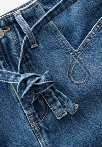 Denim tkanina s svijetloplavim pranjem, koja sadrži čvorastast vez na struku, vidljive šavove i oblikovan dizajn džepa.