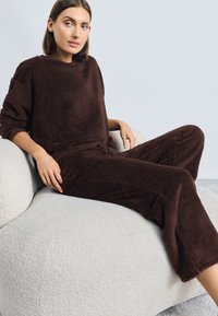 Braunes, plüschiges Loungewear-Set mit einem locker sitzenden Oberteil und Jogginghosen mit Kordelzug. Strukturiertes Material mit einer weichen, flauschigen Oberfläche. Hellgrauer Stuhl.