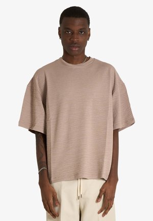 Giovane uomo che indossa una t-shirt beige testurizzata oversize e pantaloni crema chiaro con coulisse, in piedi su sfondo bianco semplice.