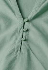 Camicia button-up verde chiaro con scollatura a V, quattro bottoni coordinati e una texture morbida, leggermente stropicciata, arricchita da dettagli cuciti.