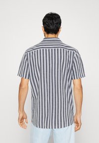 Muž stojící zády, oblečený v krátkosrsté košili s límečkem, která má vertikální šedé a bílé pruhy, a v světle modrých kalhotách, na jednobarevném pozadí.