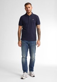 Námořnický polo tričko s límečkem, krátkým rukávem a malým logem na hrudi, v kombinaci se světle modrými džíny a bílými teniskami.