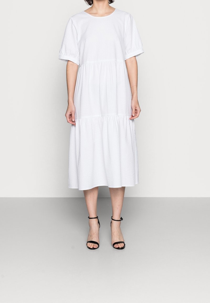 Femme portant une robe midi blanche à volants avec manches courtes et des talons noirs à bride à la cheville, debout devant un fond clair uni.