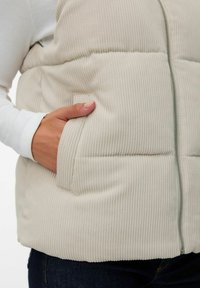 Beige corduroy puffer vest met een ritssluiting aan de voorkant, voorzien van verticale ribbelstructuur en zijzak aan de linkse heup.