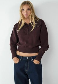 Brauner Zip-Hals-Pullover mit gerippter Textur und Cropped-Design, kombiniert mit dunkelblauen Jeanshosen mit Knopf und Taschen.