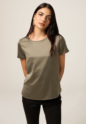 Olivgrünes kurzärmliges Blusenshirt mit glatter Textur, runder Ausschnitt und lockerer Passform. Kombiniert mit schwarzen Hosen für einen Kontrast.