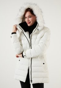 Parka blanche rembourrée avec une capuche en fausse fourrure, dotée d'une fermeture éclair, d'accents noirs, de deux poches latérales et de poignets côtelés pour plus de chaleur.