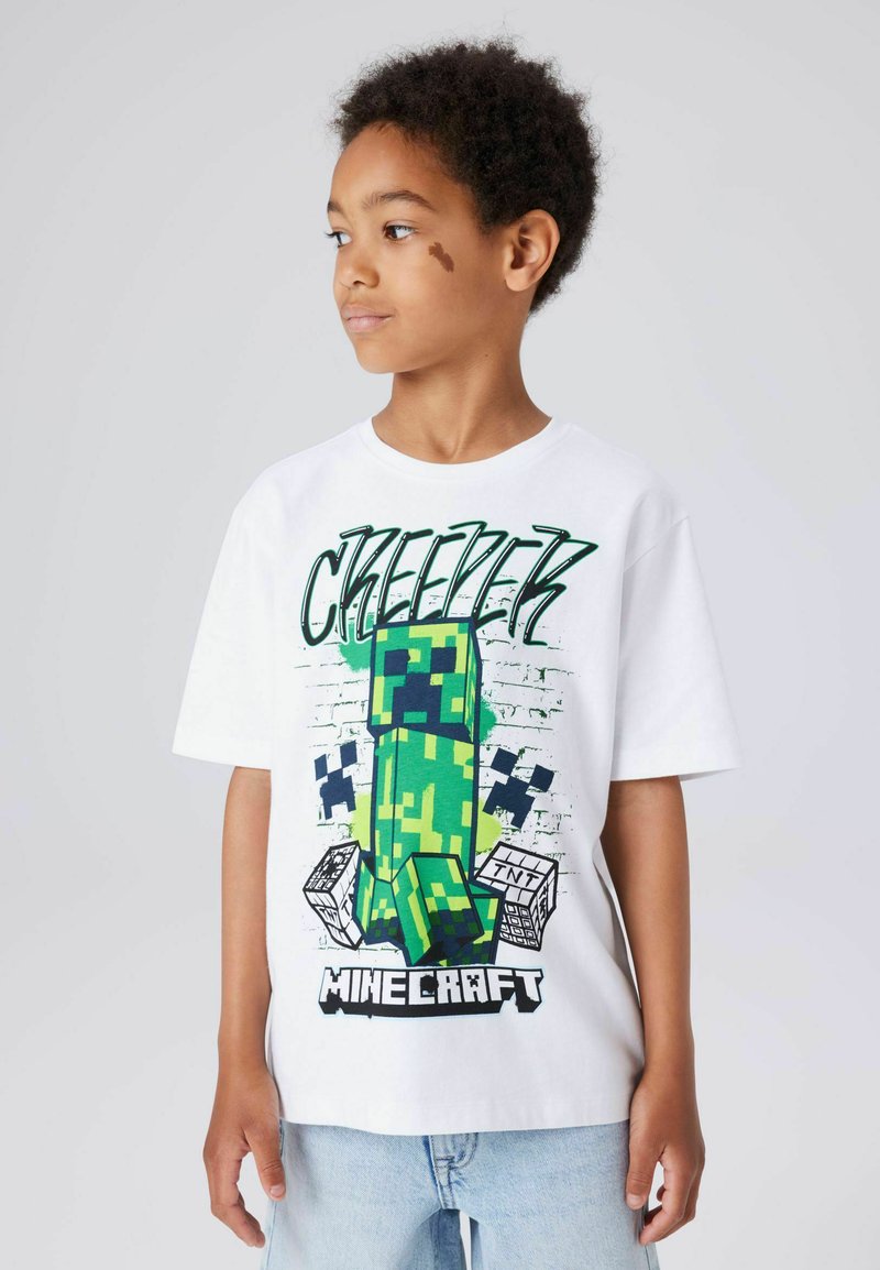 Camiseta blanca con un gráfico verde de Minecraft Creeper, con patrones pixelados y texto "Minecraft", combinada con vaqueros azul claro.