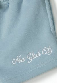 Světle modré látkové šortky s bílým vyšívaným nápisem "New York City." Měkká textura s hladkým povrchem a neformální design.