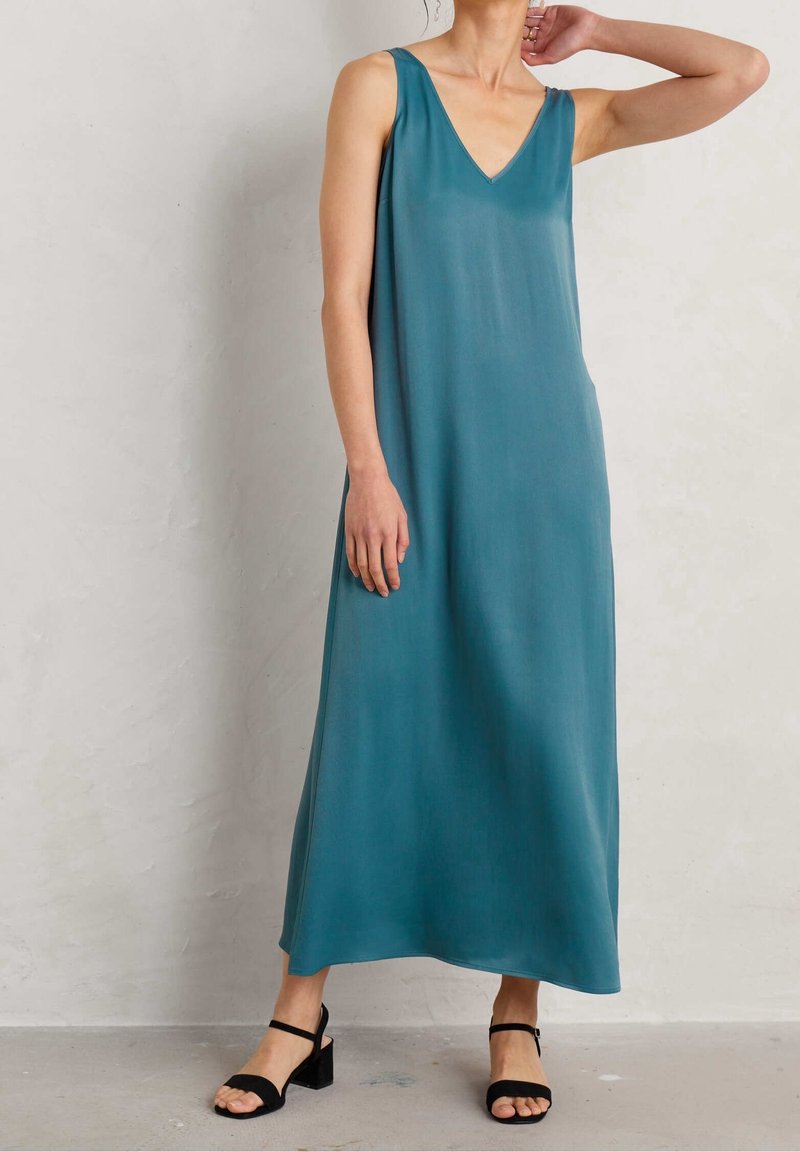 Femme portant une robe maxi sans manches de couleur sarcelle avec un décolleté en V, associée à des sandales à talons blocs noires, debout devant un mur clair uni.