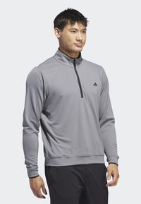 Pull-over de sport gris avec un design à demi-fermeture éclair, fabriqué à partir d'un tissu texturé, comportant des manches longues et un petit logo noir sur la poitrine.