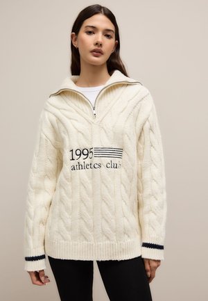 Jeune femme portant un pull en maille blanche avec le texte « 1995 athletics club » et un col zippé à moitié, debout devant un fond uni.