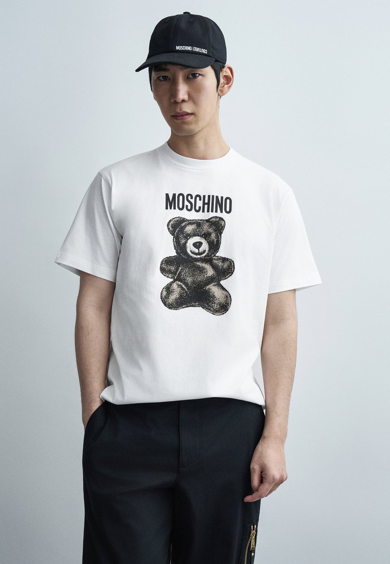 Λευκό βαμβακερό μπλουζάκι με μαύρο σχέδιο αρκουδάκι και το λογότυπο της μάρκας "Moschino" τυπωμένο πάνω. Συνδυασμένο με μαύρο παντελόνι.