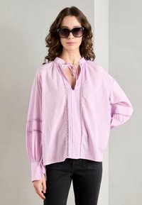 Helle rosa, übergroße Bluse mit V-Ausschnitt und Bindedetail. Mit Spitzenbesatz und gerafften Ärmeln. Getragen mit dunklen Jeans und übergroßen Sonnenbrillen.