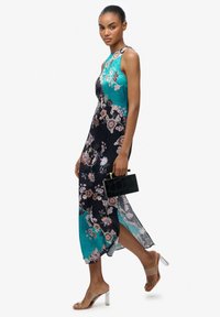 Kwiatowa sukienka maxi w turkusowym i czarnym jedwabiu, z wysokim dekoltem, bocznym rozcięciem i gładką fakturą. W połączeniu z czarną kopertówką i przezroczystymi obcasami.