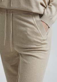 Beige jogger bikses, kas izgatavotas no mīksta auduma, ar elastīgu jostu un auklu, kā arī sānu kabatām, demonstrējot gludu tekstūru.