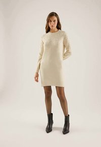 Crème gebreide sweaterjurk met lange mouwen, voorzien van decoratieve kralen, een geribde zoom, en gecombineerd met zwarte enkellaarsjes en sheer panty’s.