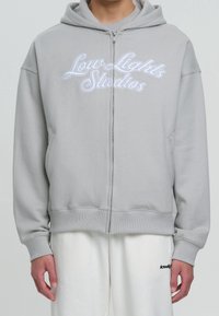Grauer Kapuzenpullover mit Reißverschluss aus weichem Stoff, mit Vorderentaschen und weiß besticktem "Low Lights Studios" Schriftzug. Klassischer Schnitt mit Kapuze.